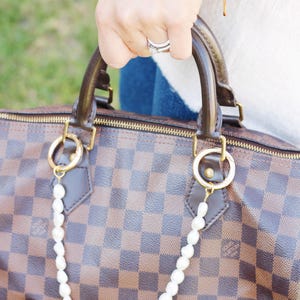 May include: A brown and tan checkered handbag with a pearl chain. The bag features brown leather handles and gold-tone hardware. The pearl chain is attached with gold rings.