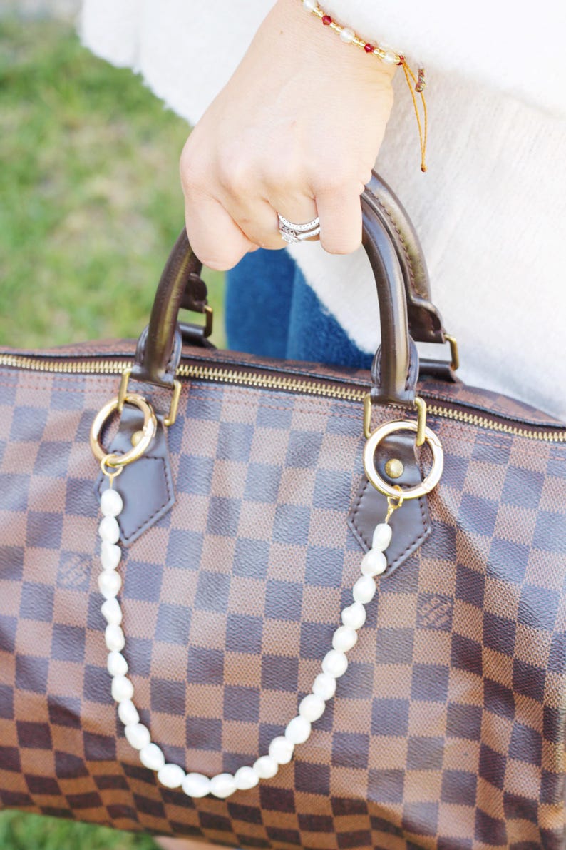 May include: A brown and tan checkered handbag with a pearl chain. The bag features brown leather handles and gold-tone hardware. The pearl chain is attached with gold rings.