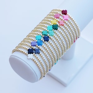 May include: A collection of gold beaded bracelets, each adorned with a colorful four-leaf clover charm. The bracelets are displayed on a white stand, showcasing a variety of colors including pink, yellow, blue, and green.