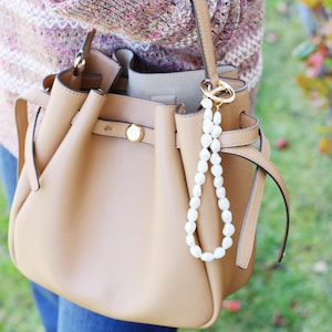 May include: A tan leather handbag with a top handle and a pearl beaded charm. The bag has a gold clasp and a matching tan strap. The person is wearing a pink and white sweater and blue jeans.