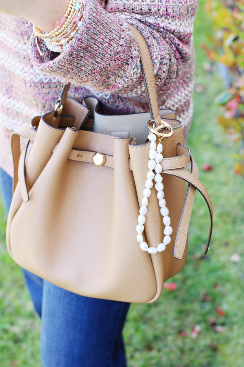 May include: A tan leather handbag with a top handle and a pearl beaded charm. The bag has a gold clasp and a matching tan strap. The person is wearing a pink and white sweater and blue jeans.