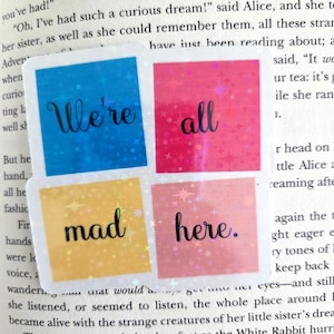 May include: A square sticker with the words "We're all mad here" in black script, each word on a different colored square. The squares are blue, pink, yellow, and pink, with a holographic, glittery effect.