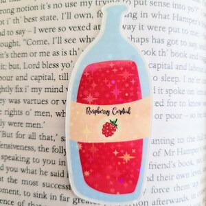May include: A sticker in the shape of a bottle, filled with red liquid and sparkling stars. The label reads "Raspberry Cordial" with a raspberry illustration. The sticker has a light blue outline.
