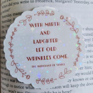 May include: A round, iridescent sticker with a scalloped edge, featuring the quote "WITH MIRTH AND LAUGHTER LET OLD WRINKLES COME" in burgundy text. The sticker has a floral border and is from "THE MERCHANT OF VENICE."