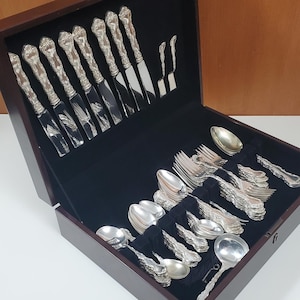May include: An open, dark wooden box containing a complete set of silver cutlery. The set includes knives, forks, and spoons, all with decorative handles. The cutlery is arranged on a black velvet surface.