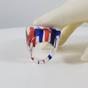 Peut inclure: Une bague triangulaire transparente avec des rayures rouges, blanches et bleues. La bague est en plastique et a un design géométrique unique.