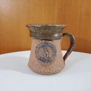May include: A brown and tan ceramic mug with a brown glaze. The mug has a handle and a circular design on the front that says "The Island Penguin" with a penguin inside the circle.
