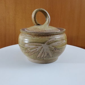 Peut inclure: Un pot en céramique avec couvercle, de couleur brun et beige. Le pot a une poignée unique et un motif de feuille stylisé.