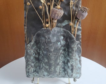 May include: A rectangular ceramic wall art piece with a dark grey background and a turquoise-coloured lower section. Dried wheat, seed pods, and other dried plants are arranged in the centre. The piece is displayed on a small metal stand.