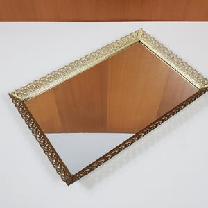 Peut inclure: Un plateau de vanité rectangulaire avec un cadre en filigrane doré. La surface du miroir reflète un fond en bois. Les bords du plateau présentent un design orné et détaillé, ajoutant une esthétique vintage. Le plateau est incliné, mettant en valeur ses éléments décoratifs.