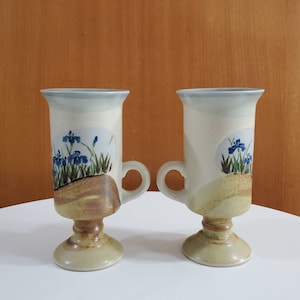 May include: Two ceramic mugs with a pedestal base. The mugs are decorated with a blue and green floral design on a white background. The base is a light brown colour.