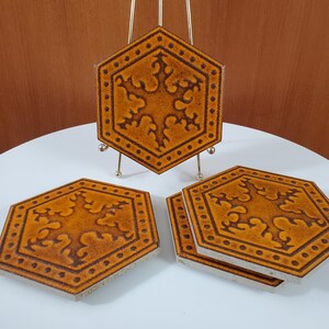 May include: Set of four hexagonal ceramic coasters. The coasters are a warm orange colour with a dark brown decorative pattern. One coaster is displayed on a small gold stand. The coasters are approximately 10 cm in diameter.