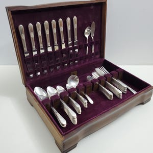 May include: A wooden box with a purple velvet interior contains a set of silver cutlery. The cutlery includes forks, knives, and spoons.