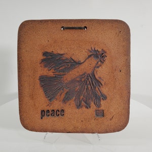 May include: A brown ceramic wall hanging with a stylized bird design and the word "peace" inscribed below. The bird is in flight with its wings spread wide.