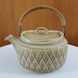 May include: A ceramic teapot with a light brown glaze and a woven wicker handle. The teapot has a leaf pattern embossed on the body.