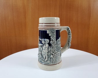May include: A ceramic beer stein with a blue and white design. The stein features a raised design of a man holding a beer mug and a building with a vine growing on it. The stein has a brown handle.