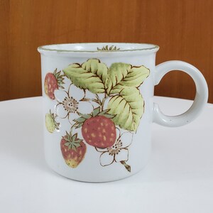 Peut inclure: Une tasse en céramique blanche avec un motif de fraises brun, vert et rose. La tasse a une poignée arrondie.