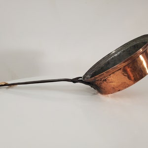 Peut inclure: Une casserole en cuivre vintage avec une longue poignée en bois brun foncé. La casserole a un fond arrondi et un bord légèrement évasé.