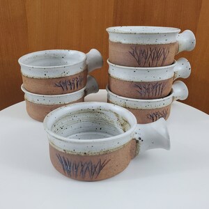 May include: A stack of six white ceramic soup bowls with brown speckled glaze and a brown band around the top. Each bowl has a brown handle and a design of brown grass on the brown band.