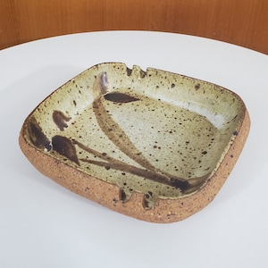 May include: A square ceramic ashtray with a speckled brown and white glaze. The ashtray has a brown design that resembles a stylized flower.