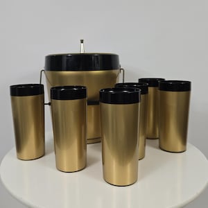 May include: A gold and black ice bucket with six matching gold and black tumblers. The ice bucket has a lid with a small spout.