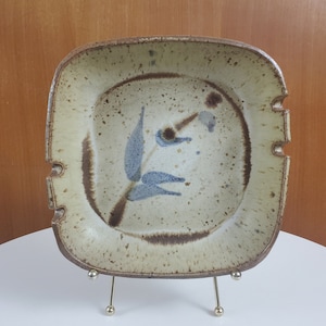 May include: Square ceramic dish with a speckled beige surface and a brown rim. The dish features a blue leaf design and two cut-out notches on opposite sides. The dish is displayed on a gold-colored metal stand.