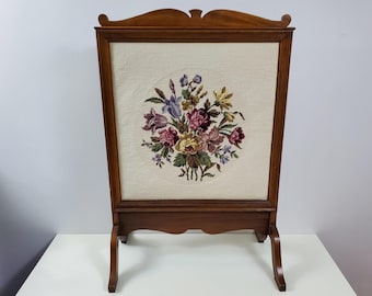 Può includere: Uno schermo per camino in legno con un pannello ricamato con fiori. Lo schermo presenta un bouquet di fiori rosa, viola e gialli su sfondo bianco.