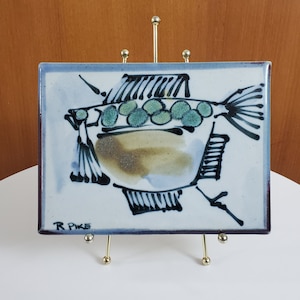 May include: A white ceramic tile with a black and white illustration of a fish. The fish has green spots and the tile is signed "R Pike".
