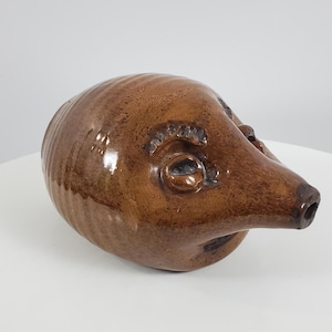 May include: A brown ceramic pitcher shaped like a pig's head with a spout. The pitcher has a glazed finish and features a detailed face with eyes, a snout, and a textured surface.