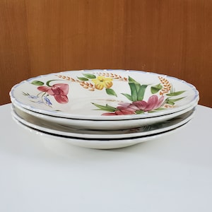 May include: Three white ceramic plates stacked on top of each other, each with a floral pattern. The plates have a scalloped edge and a blue trim. The floral design features red, yellow, and blue flowers.