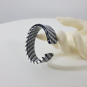 May include: A black and white patterned cuff bracelet. The bracelet has a curved, open design and is made of a glossy material. The pattern consists of thin, diagonal lines creating a woven effect. The bracelet is displayed on a white surface.