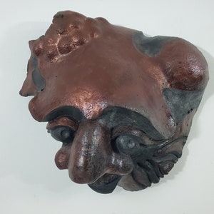 May include: A ceramic sculpture of a face with a textured surface. The face is brown and black with a large nose and exaggerated features.