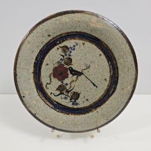 May include: A ceramic plate with a brown, blue and white floral design featuring a bird. The plate has a speckled glaze and a brown rim.