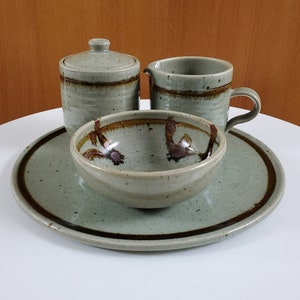 May include: A green ceramic serving set with a brown rim. The set includes a small bowl, a creamer, and a sugar bowl with a lid. The set is on a white surface.