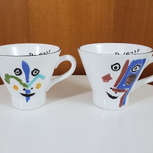 May include: Two white ceramic mugs with abstract face designs. One mug has a blue, green, and yellow face design. The other mug has a blue, red, and brown face design. Both mugs have black writing on the rim.