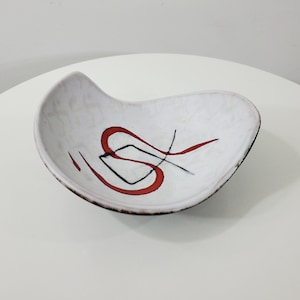 May include: White ceramic bowl with a red and black abstract design. The bowl has a unique, asymmetrical shape.