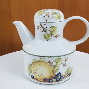 May include: White ceramic teapot with a floral design featuring white flowers, green leaves, and a yellow lemon with blue berries. The teapot has a lid with a floral design and a handle.
