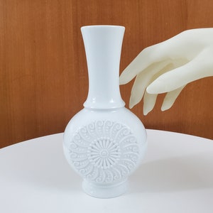 May include: White ceramic vase with an embossed floral design. The vase has a round base and a narrow neck.