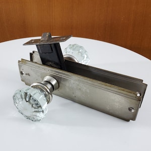 Peut inclure: Quincaillerie de porte antique avec une finition argentée et deux boutons en verre transparent. La quincaillerie a une plaque rectangulaire avec un bord décoratif et un mécanisme de loquet.