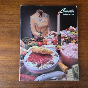 May include: Cover of "Ceramics Monthly" magazine, December 1969, featuring a still life with a ceramic shirt, food, and a lit candle. The image has a vintage aesthetic with vibrant colors and a focus on ceramic art.