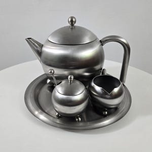 May include: A brushed metal tea set comprising a teapot, creamer, sugar bowl with a spoon, and a serving tray. The teapot features a curved handle and a lid with a small knob. The set has a minimalist design.