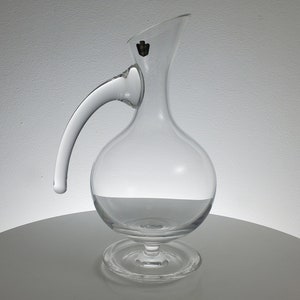 May include: A clear glass decanter with a round body and a long, curved handle. The decanter has a small, round base.