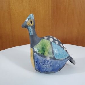 Peut inclure: Figurine d'oiseau en céramique avec un motif patchwork. L'oiseau présente un corps gris avec des accents bleus, verts et jaunes. Des pois blancs ornent le dos. L'oiseau a un long cou et un bec pointu, et il est posé sur une surface blanche.