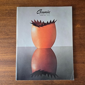 May include: The cover of Ceramics Monthly magazine, January 1983 issue, features an orange ceramic vase with a spiky top. The vase is reflected in a mirrored surface, creating a symmetrical image. The magazine is on a wooden surface.