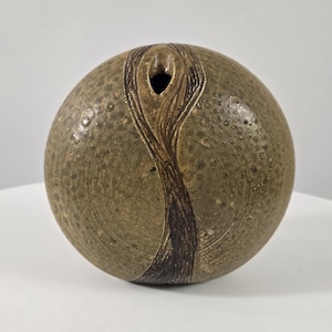 May include: A round, ceramic vase with a textured, speckled surface in shades of brown and tan. A dark brown, vertical design flows from the top opening down the front. The vase has a small opening at the top.