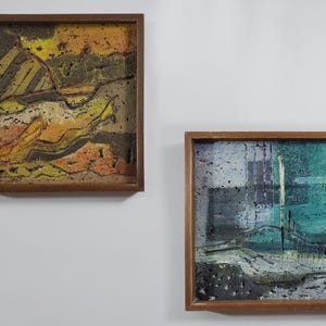 May include: Two framed abstract paintings with textured surfaces. The painting on the left has a yellow, orange, and brown color scheme. The painting on the right has a blue, green, and gray color scheme.