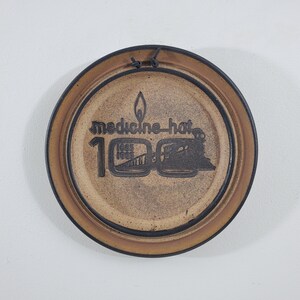 May include: A brown ceramic plate with a black rim and a black design of a train and the text "Medicine Hat 100" with a flame above the text.