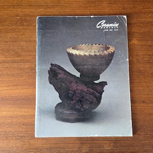 May include: A magazine cover featuring a ceramic sculpture. The sculpture is a dark brown bowl with a textured rim, supported by a dark purple, organic-shaped base. The magazine title is "Ceramics Monthly."
