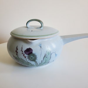 Peut inclure: Une théière en céramique blanche avec une poignée et un couvercle verts. La théière est décorée d'un motif floral avec un chardon violet.