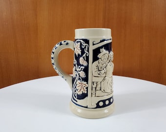 May include: A ceramic beer stein with a blue and white design. The stein features a raised relief of two people sitting on a bench. The stein has a handle and a wide base.